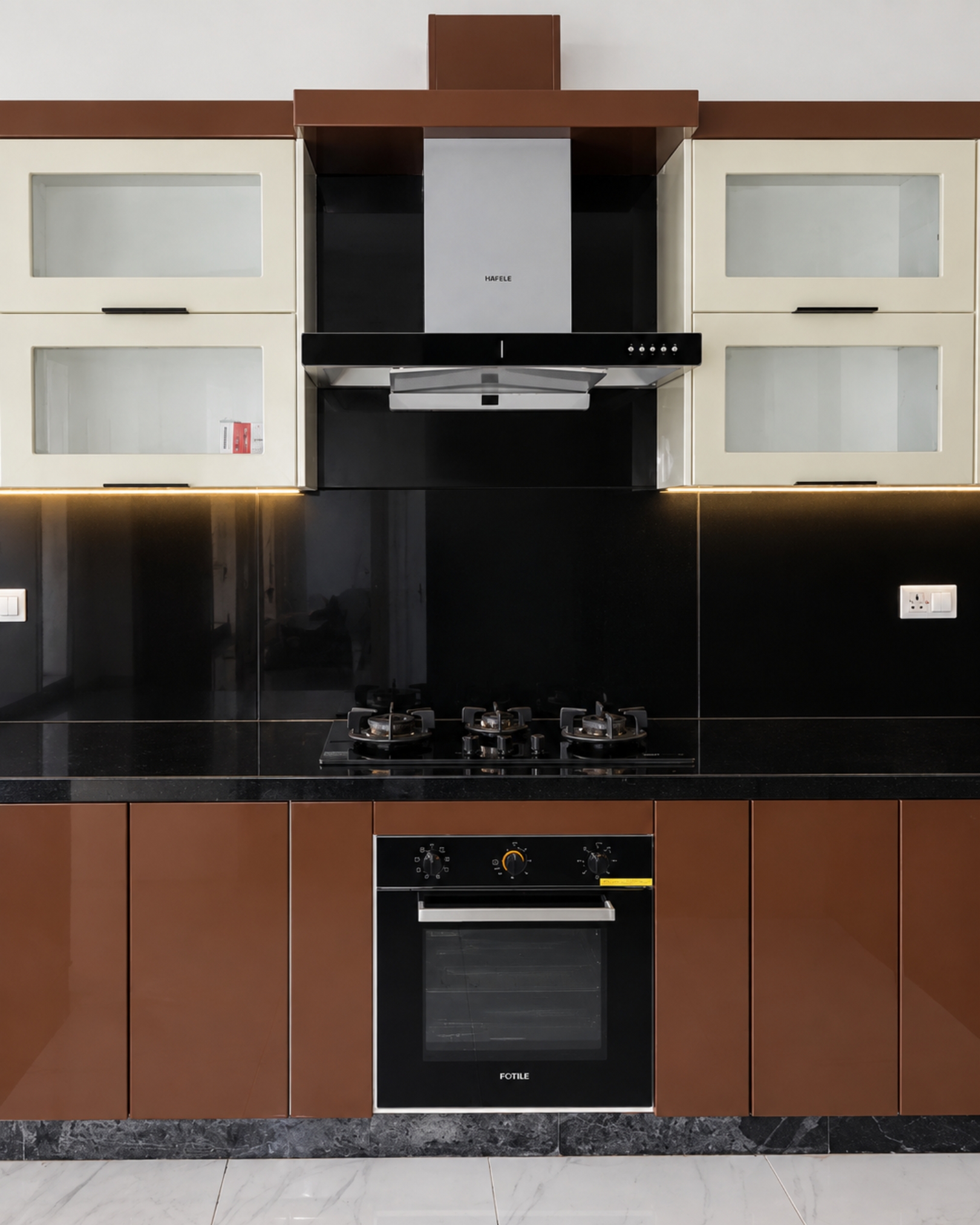 Front view of Mini Cucine’s brown and cream kitchen with glass front cabinets, a chimney hood, gas hob, built in oven and warm lighting.