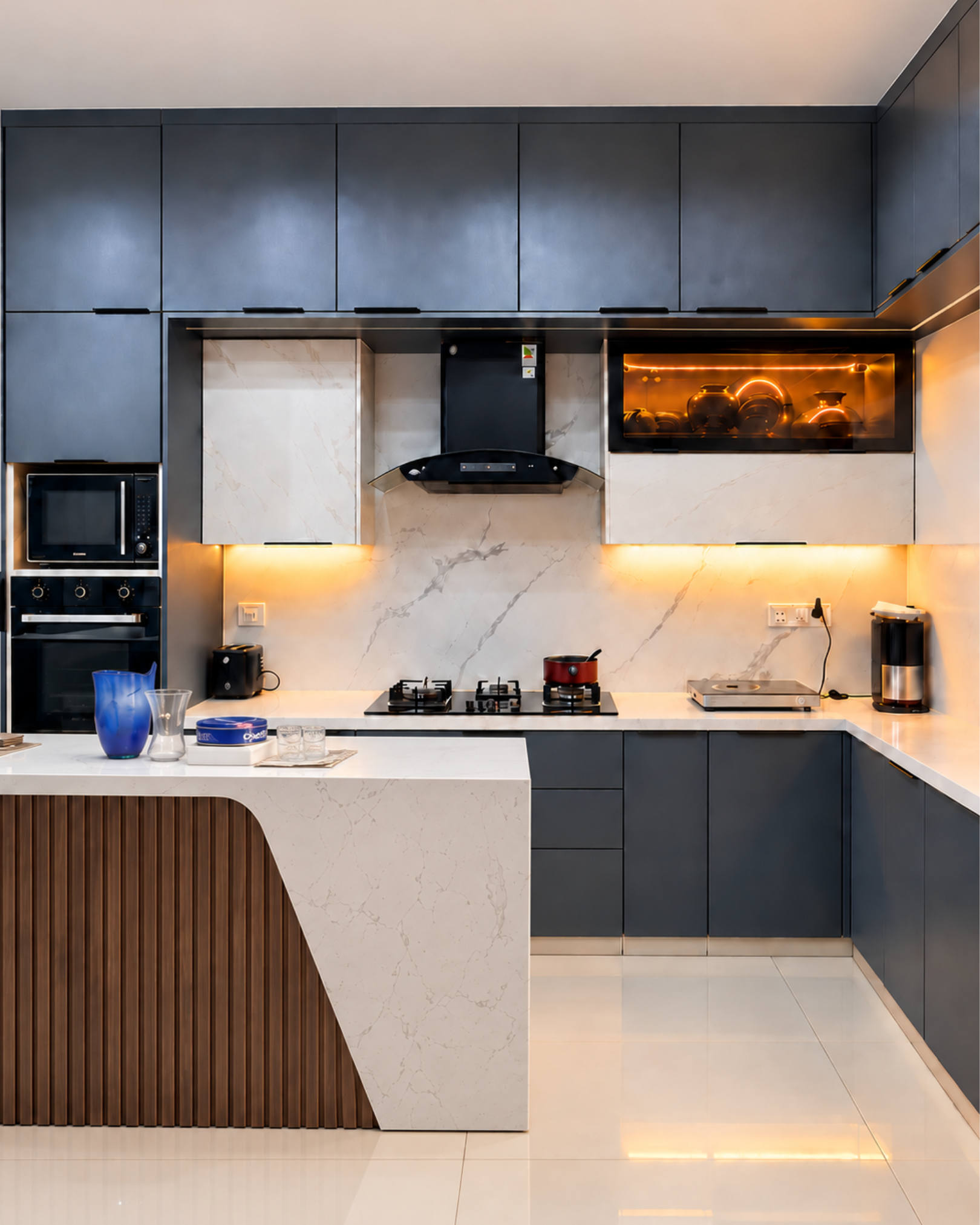 Front view of matte charcoal kitchen cabinets, built in appliances and a marble effect island with curved wood slat detailing.