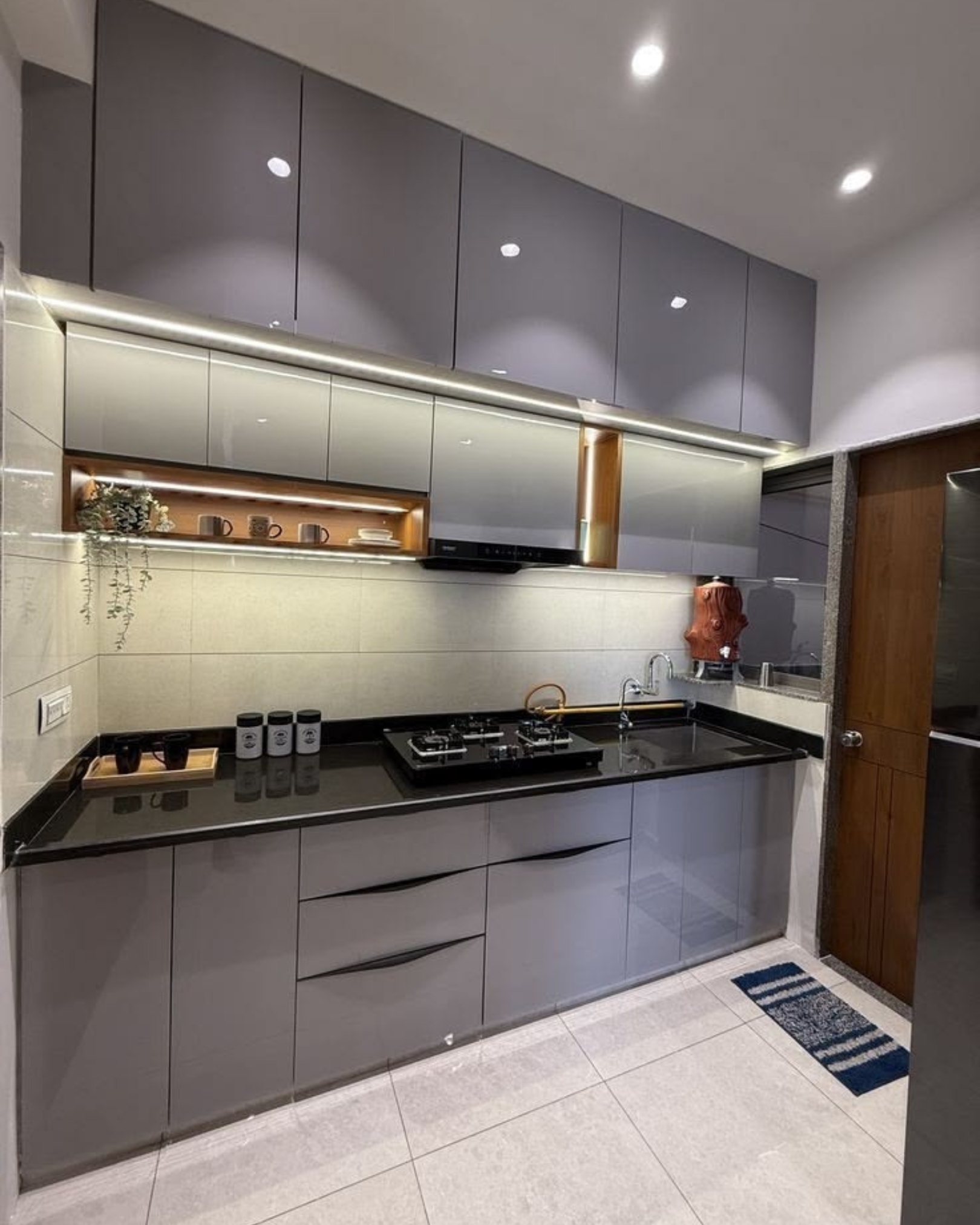Compact kitchen with glossy grey cabinets, a black countertop, illuminated wood-look shelves and a tiled backsplash.
