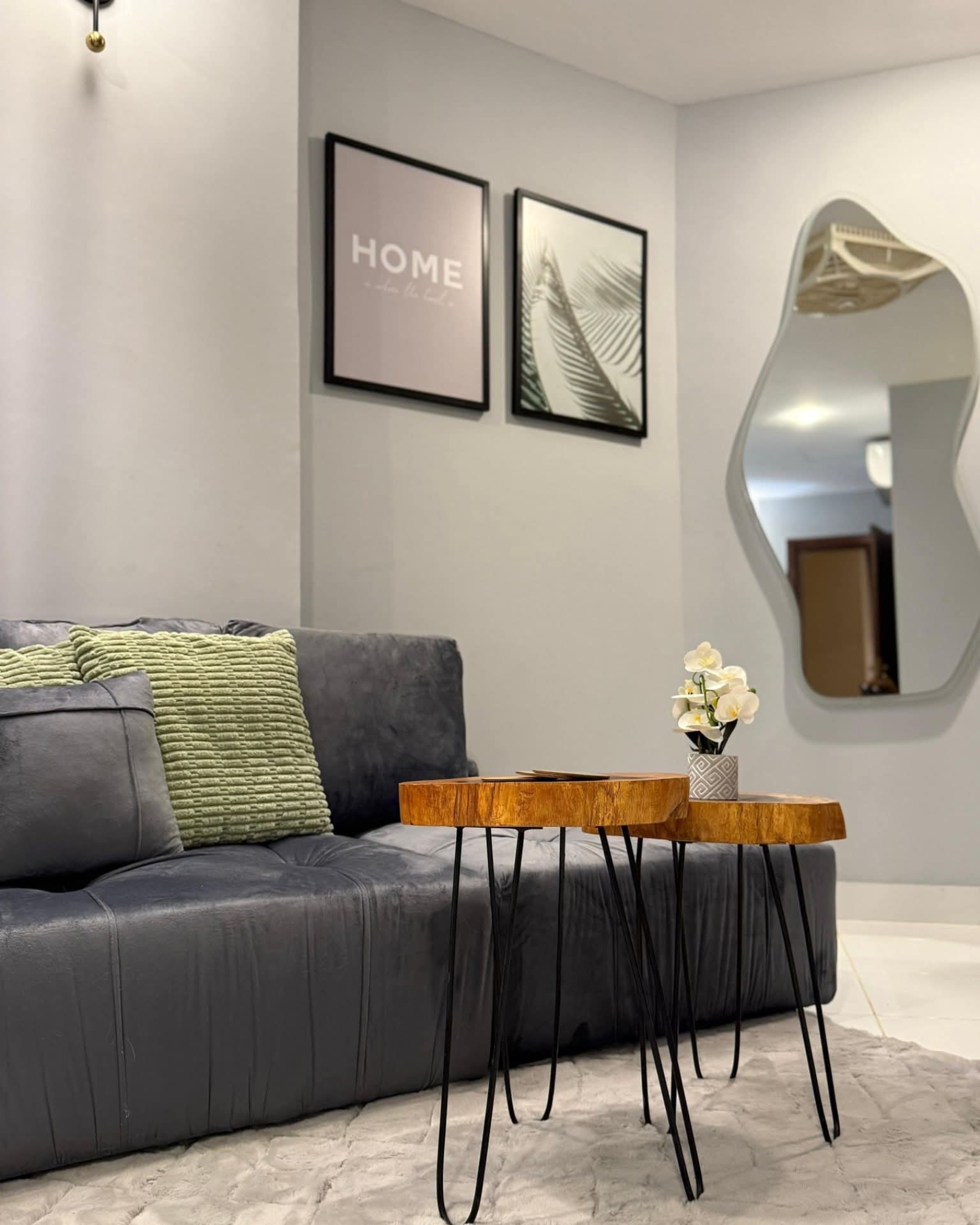 2 Live edge wooden tables with black hairpin legs beside a grey sofa, textured green cushions and an irregular wall mirror.