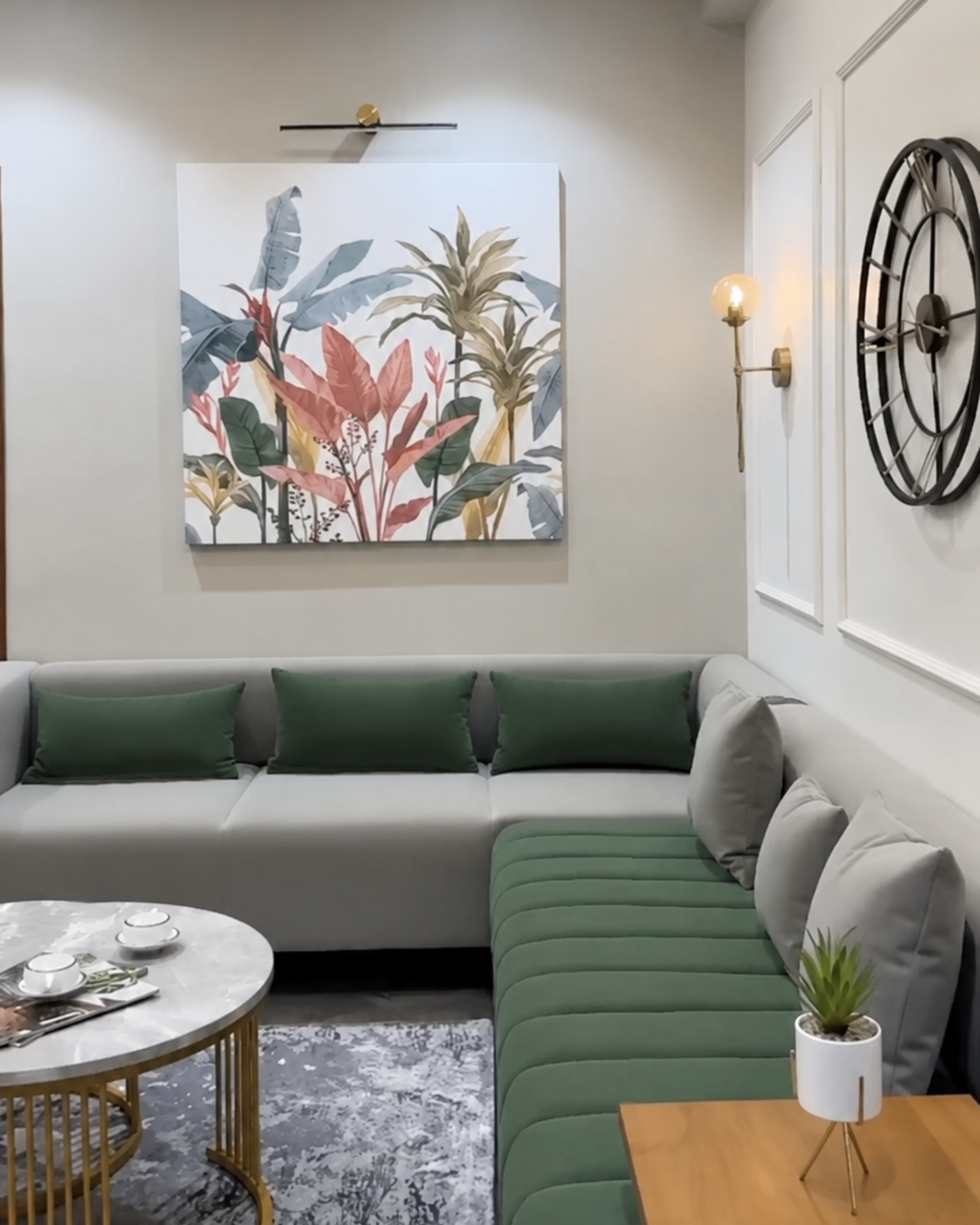 2 Grey and green corner sofa beneath colorful botanical artwork, beside a panelled wall with a clock and gold wall light.