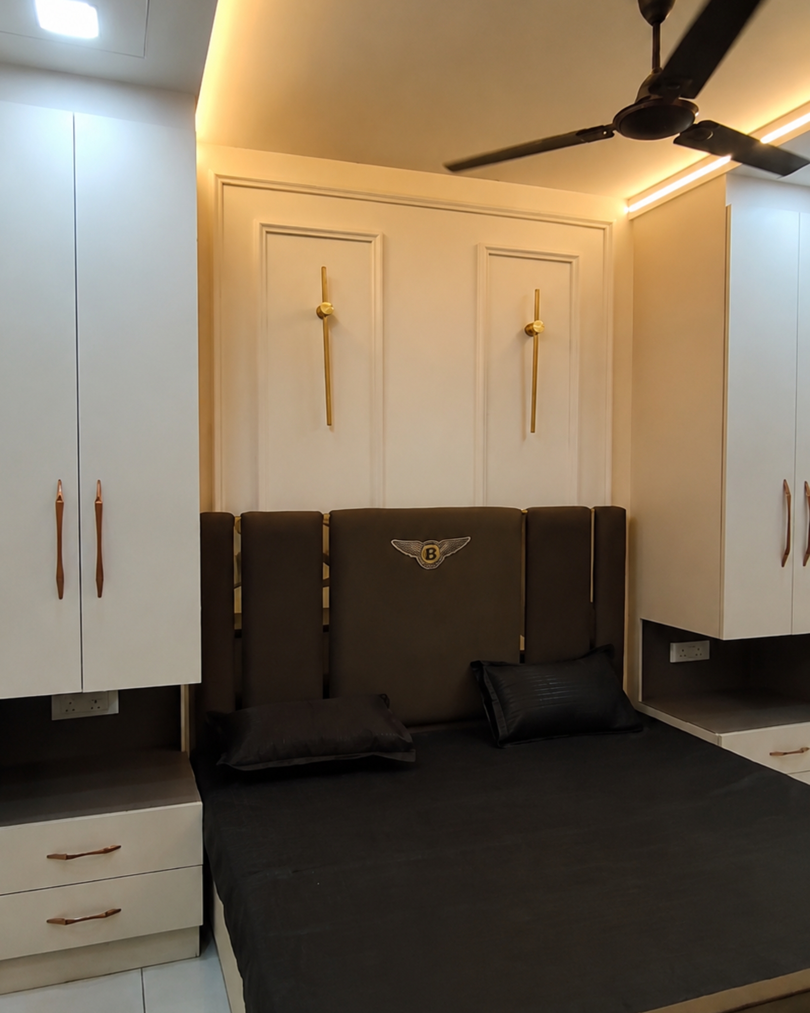 Dark upholstered bed between tall cream cabinets, with bedside drawers, gold wall lights and warm ceiling lighting.