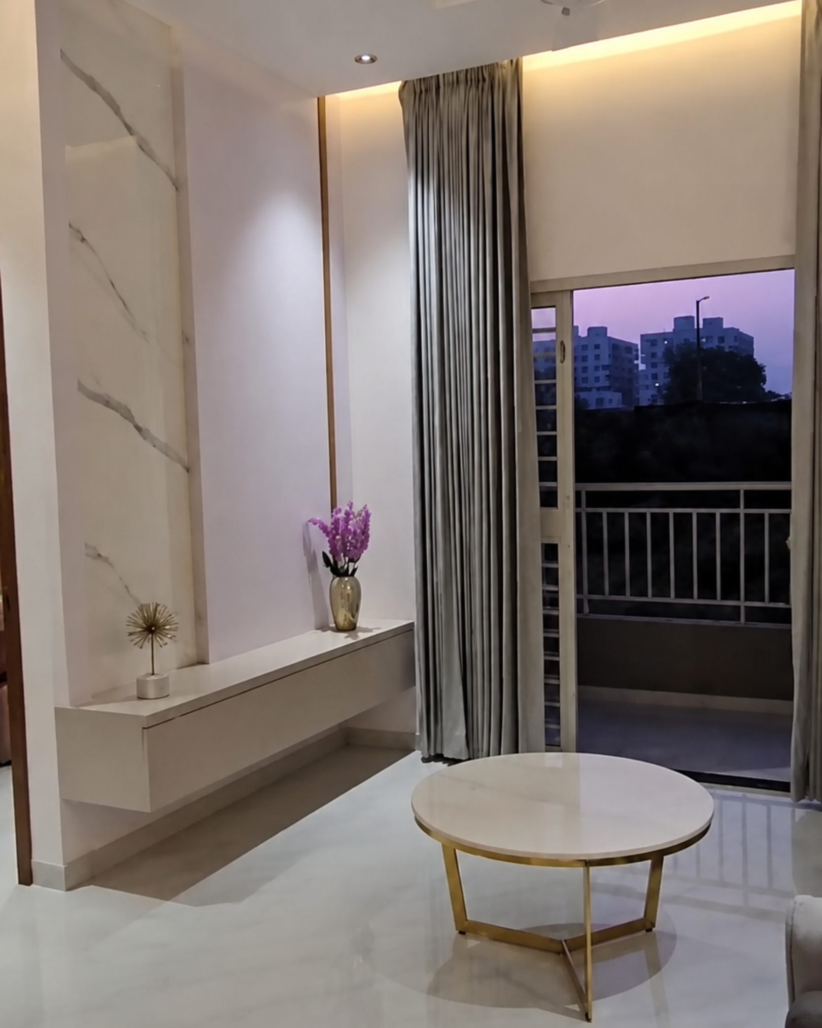 Floating cream console beneath marble effect wall detailing, with a round gold framed coffee table and full length grey curtains.