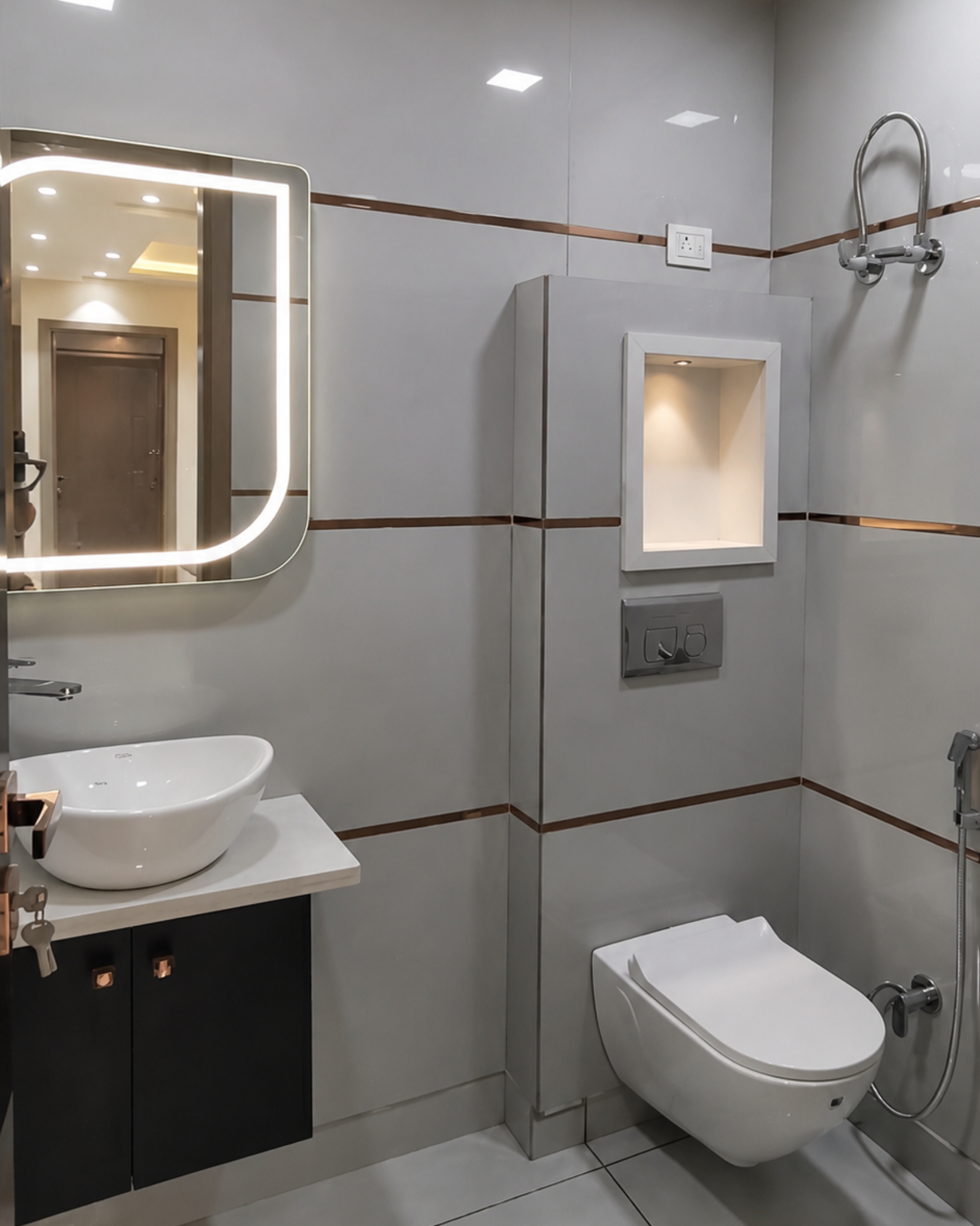Grey tiled bathroom with metallic trim, an illuminated rectangular mirror, white vessel basin and wall hung toilet.