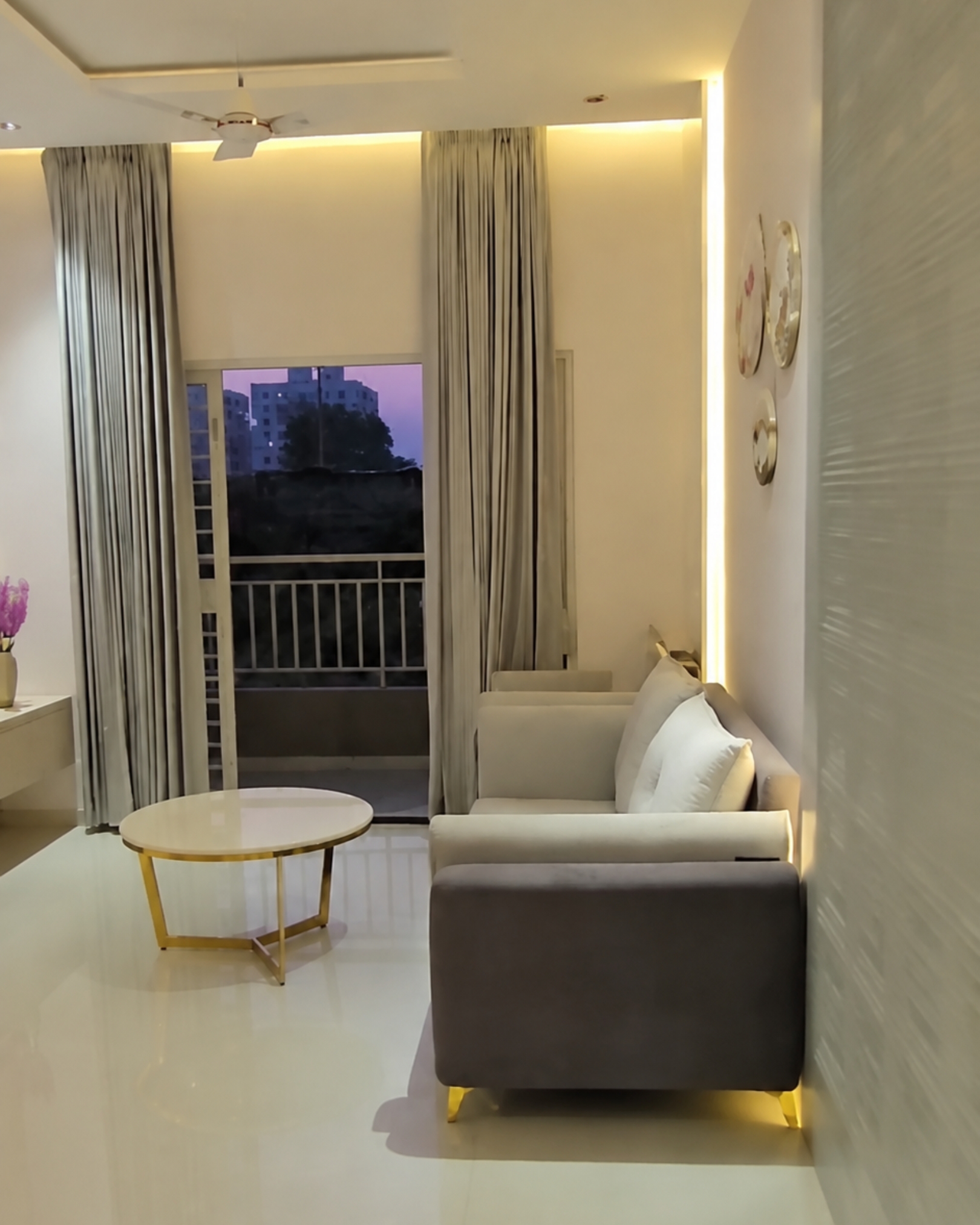 Cream and grey upholstered seating beside a round coffee table, with warm accent lighting and curtains framing the balcony door.