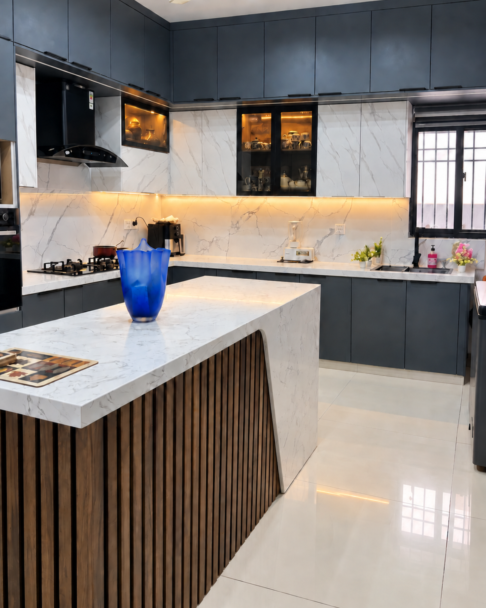 Wood slat island with a marble effect waterfall countertop and blue vase, overlooking warmly lit kitchen cabinets.
