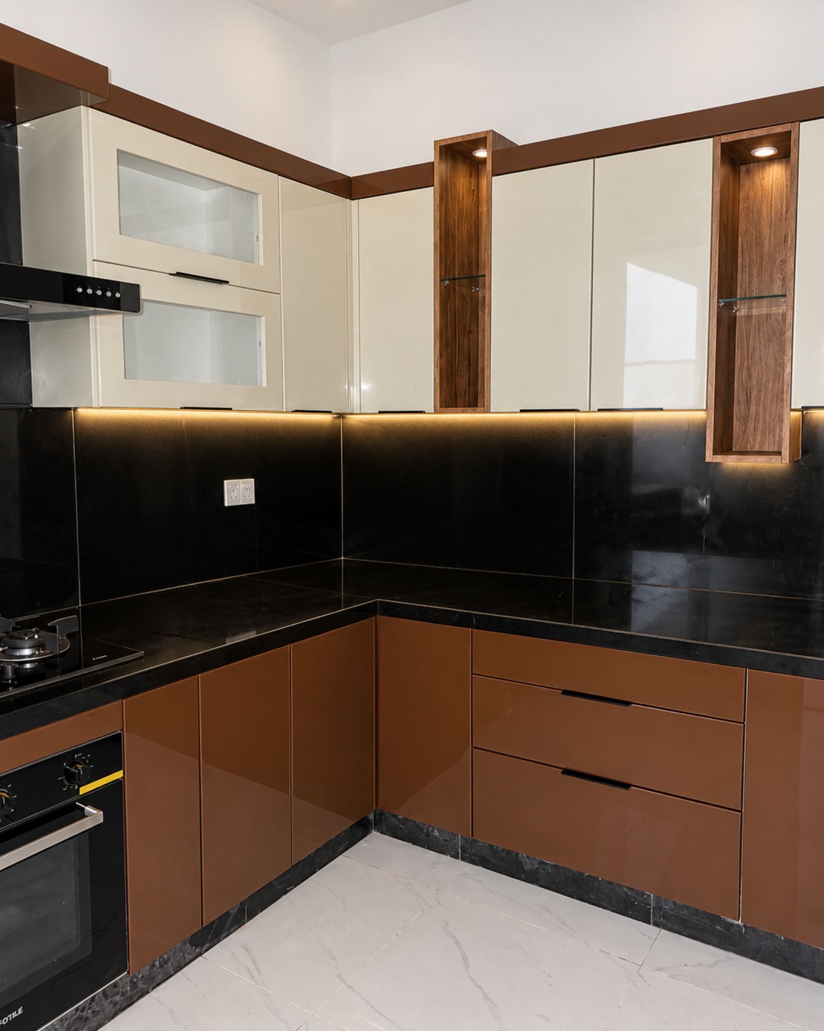 Kitchen corner featuring glossy brown drawers, cream upper cabinets, two illuminated wood-look display niches and a glossy black backsplash.