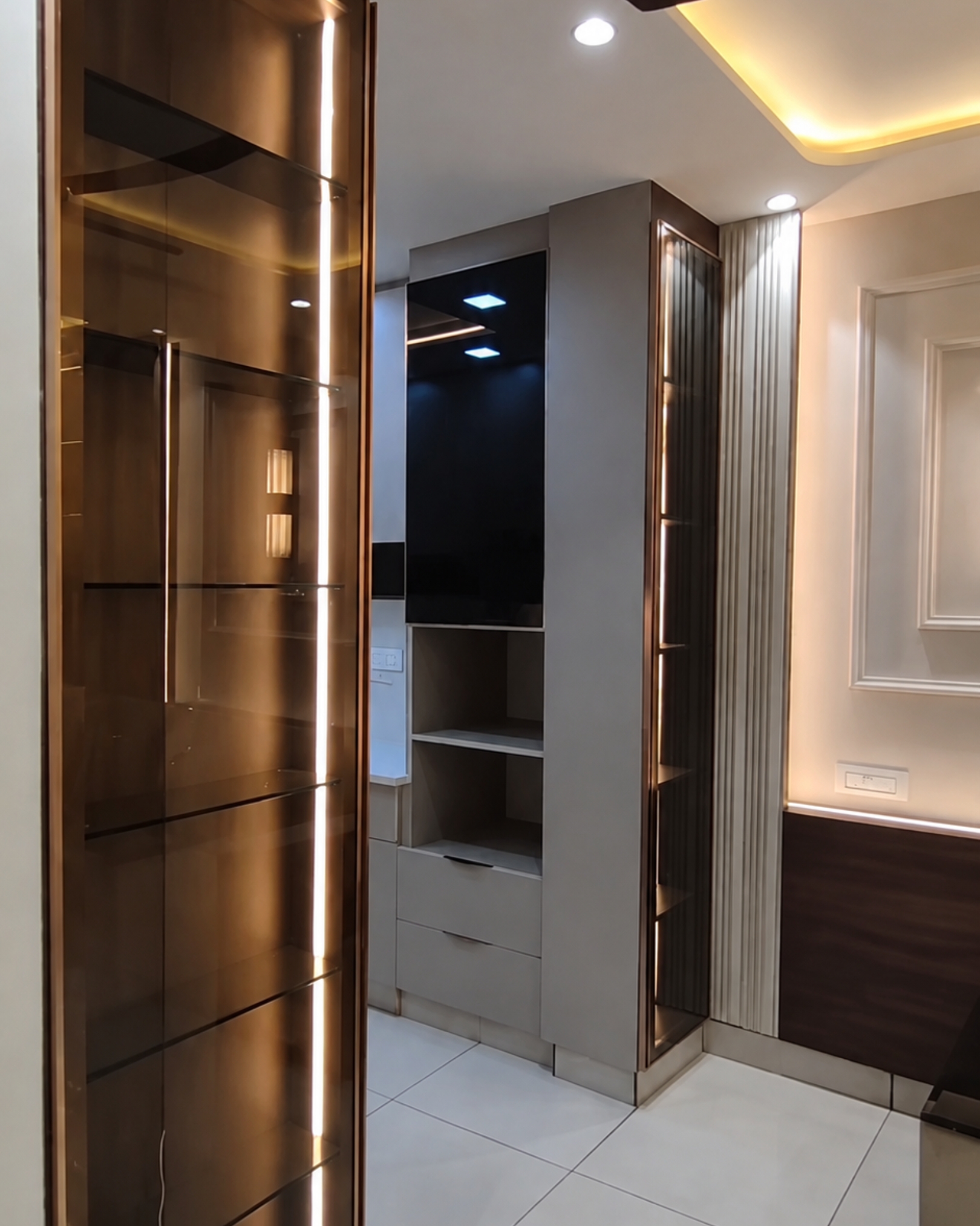 Tall glass display cabinets with bronze toned frames and vertical lighting beside grey storage units and fluted panels.