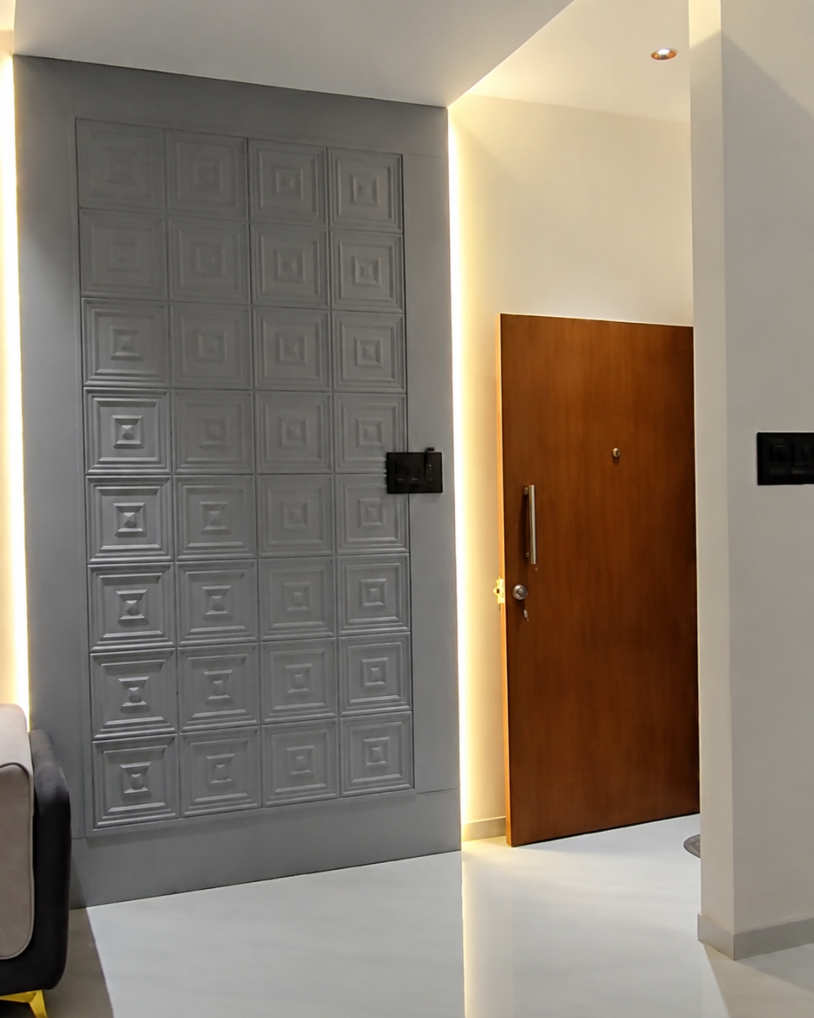 Grey geometric feature wall with raised square patterns and warm vertical lighting beside a wooden entrance door.