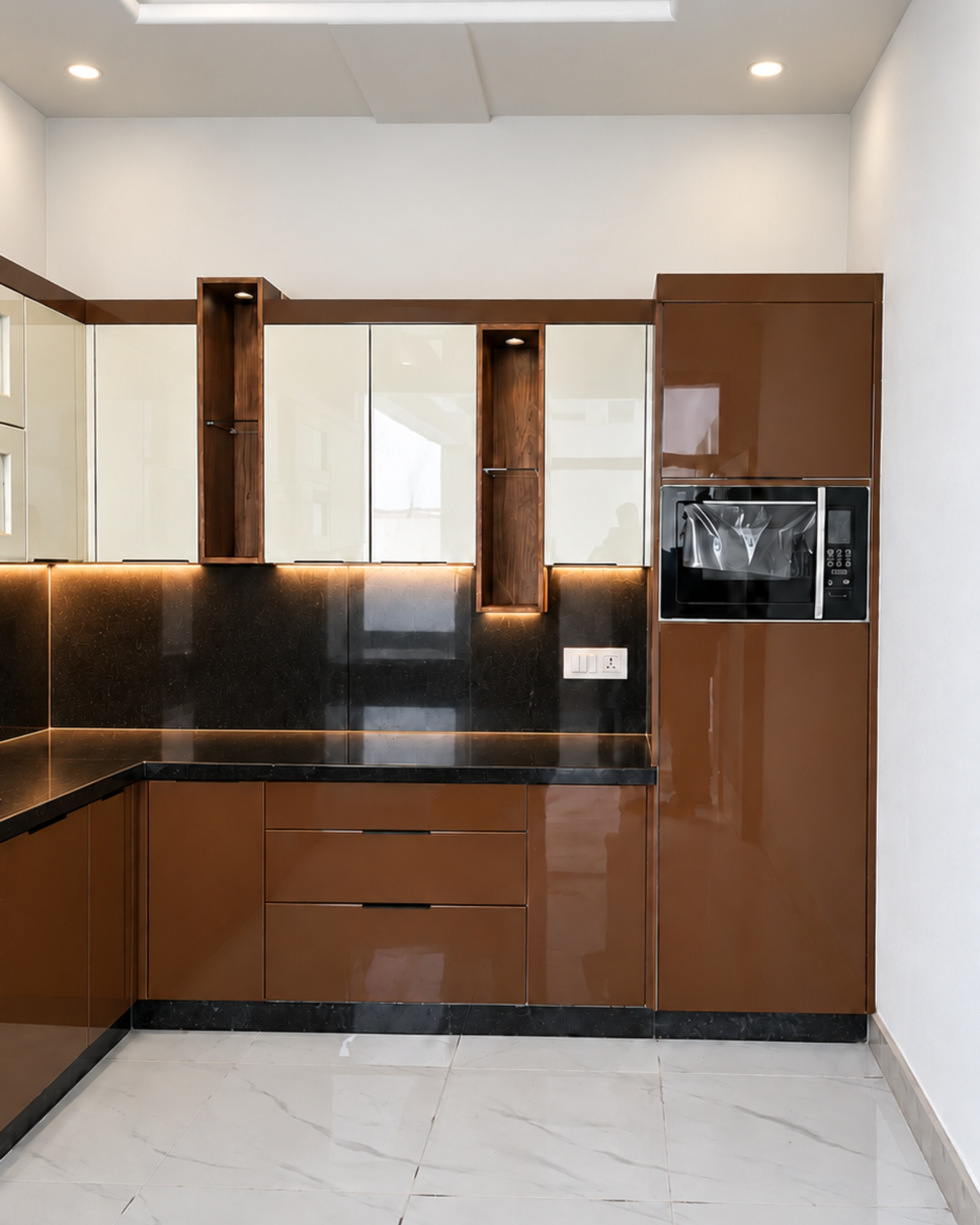 Tall brown microwave cabinet beside cream wall cabinets, illuminated display niches and three brown drawers beneath a black countertop.