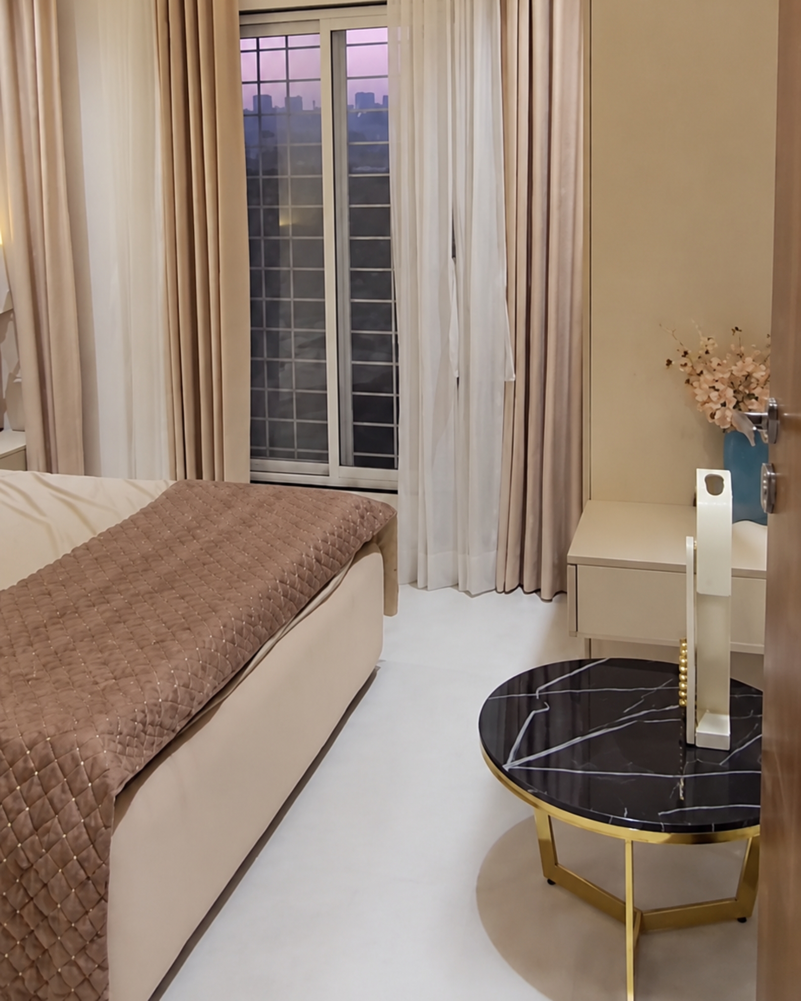 Neutral bedroom with a quilted pink throw, layered curtains, a white dressing desk and a black marble effect side table.