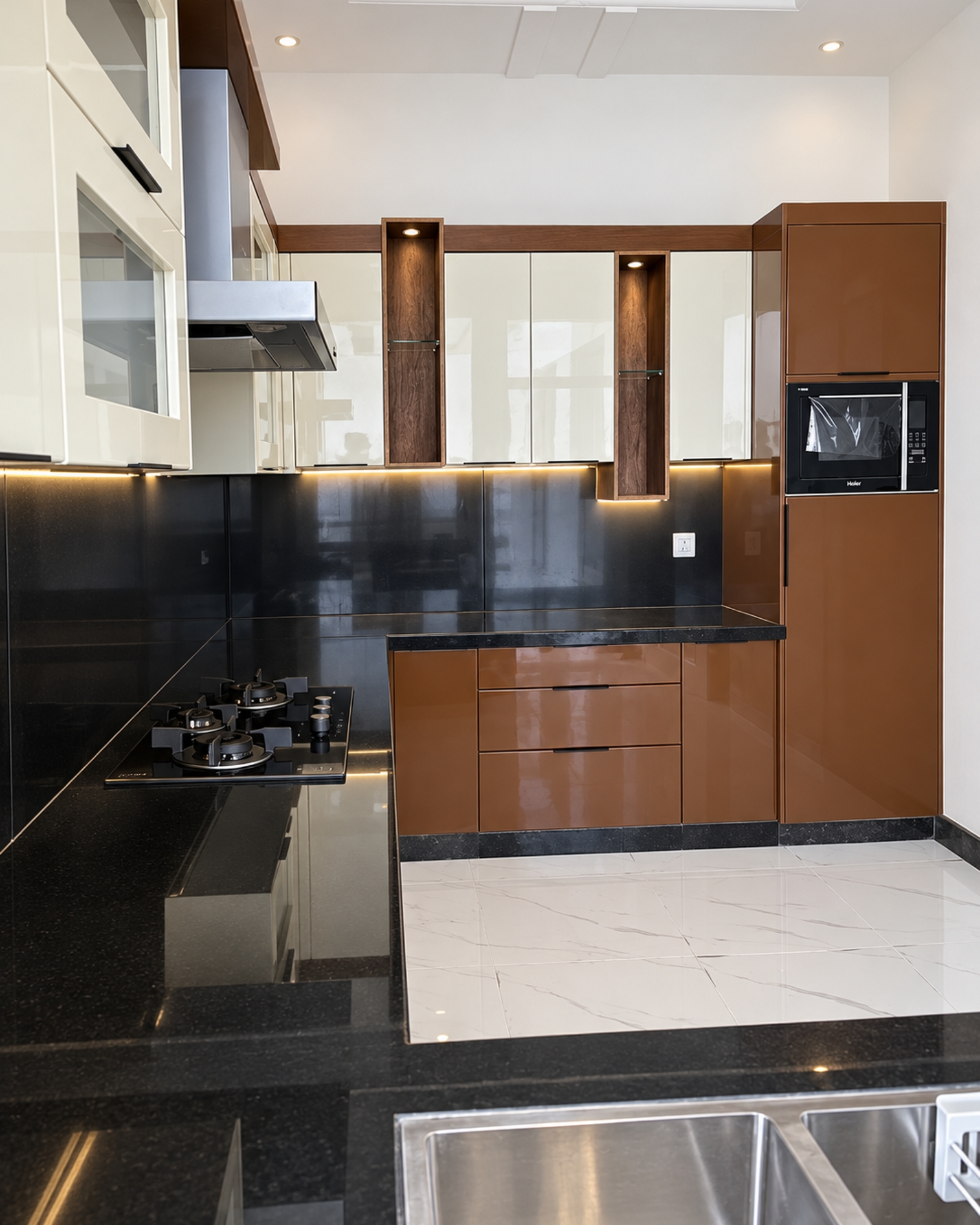 View across a double sink towards brown and cream kitchen cabinets, a gas hob, tall microwave cabinet and warm lighting.