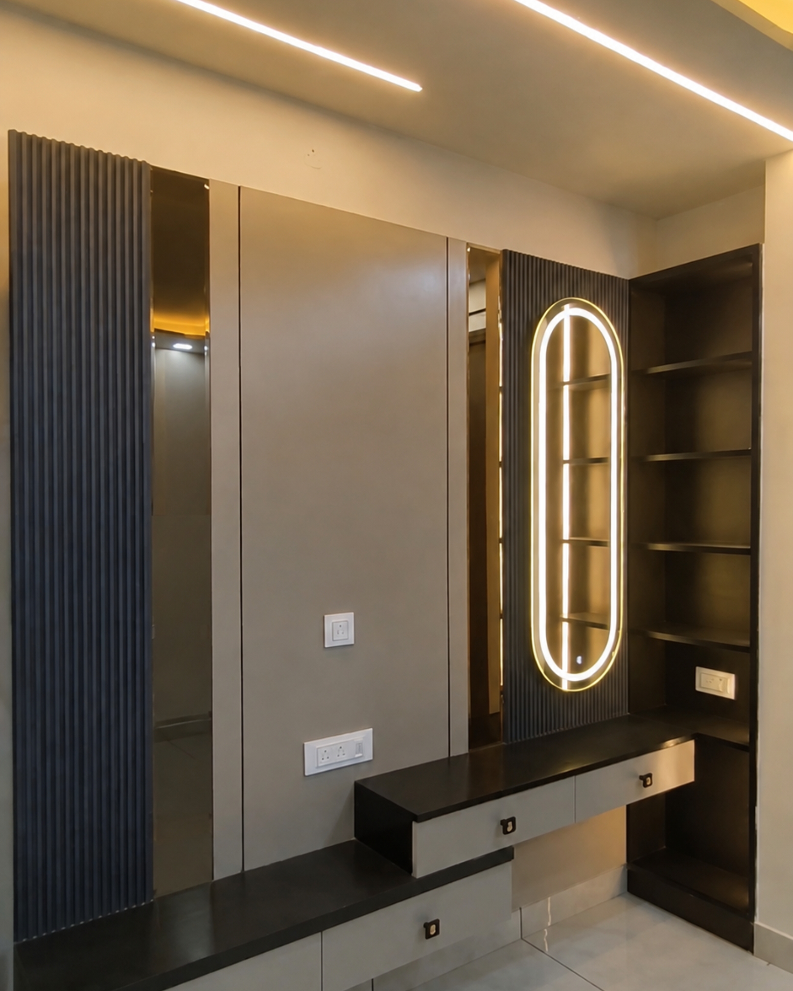 Dressing unit with an illuminated oval mirror, grey drawers, black countertop, open shelves and dark fluted wall panels.