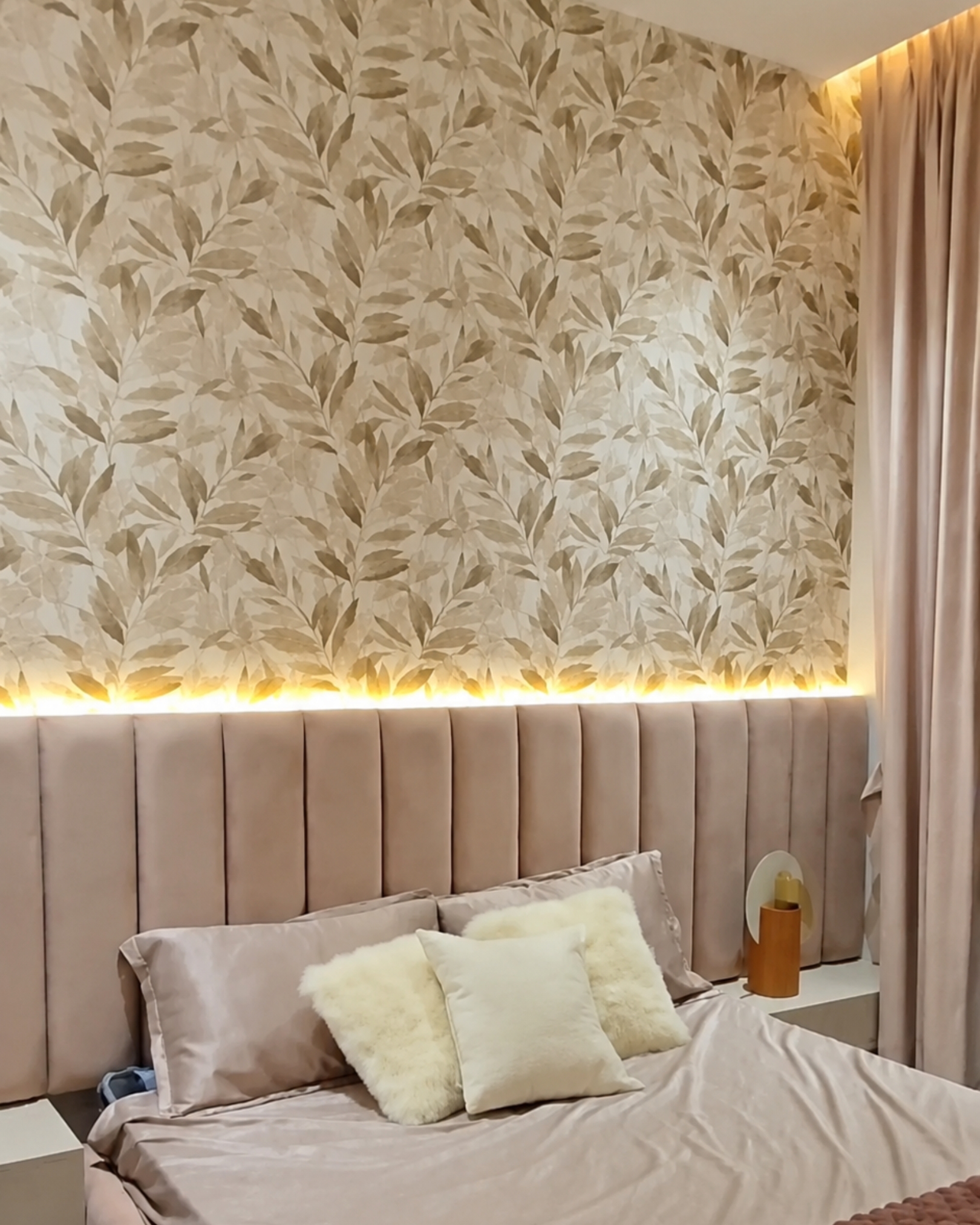 Soft pink channeled headboard beneath a beige leaf-pattern feature wall, with warm backlighting and cream cushions.