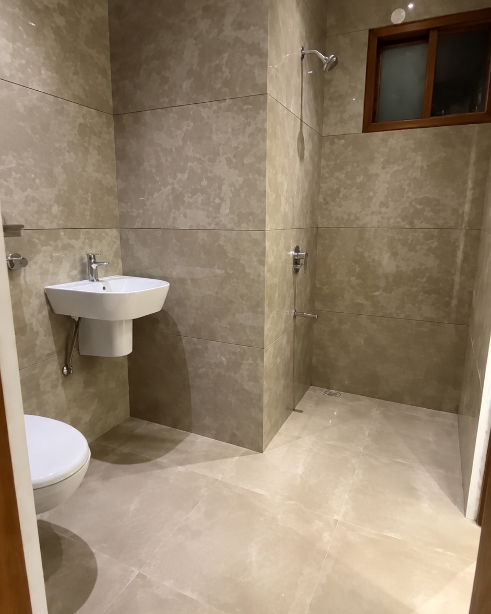 7 Beige tiled bathroom with a wall mounted white basin, chrome fittings and an open shower area beneath a small window.