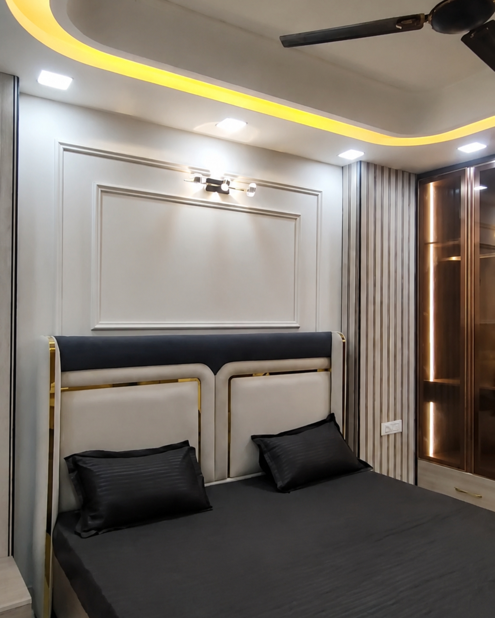 Cream and charcoal upholstered headboard with gold trim beneath wall mouldings, beside an illuminated glass cabinet.