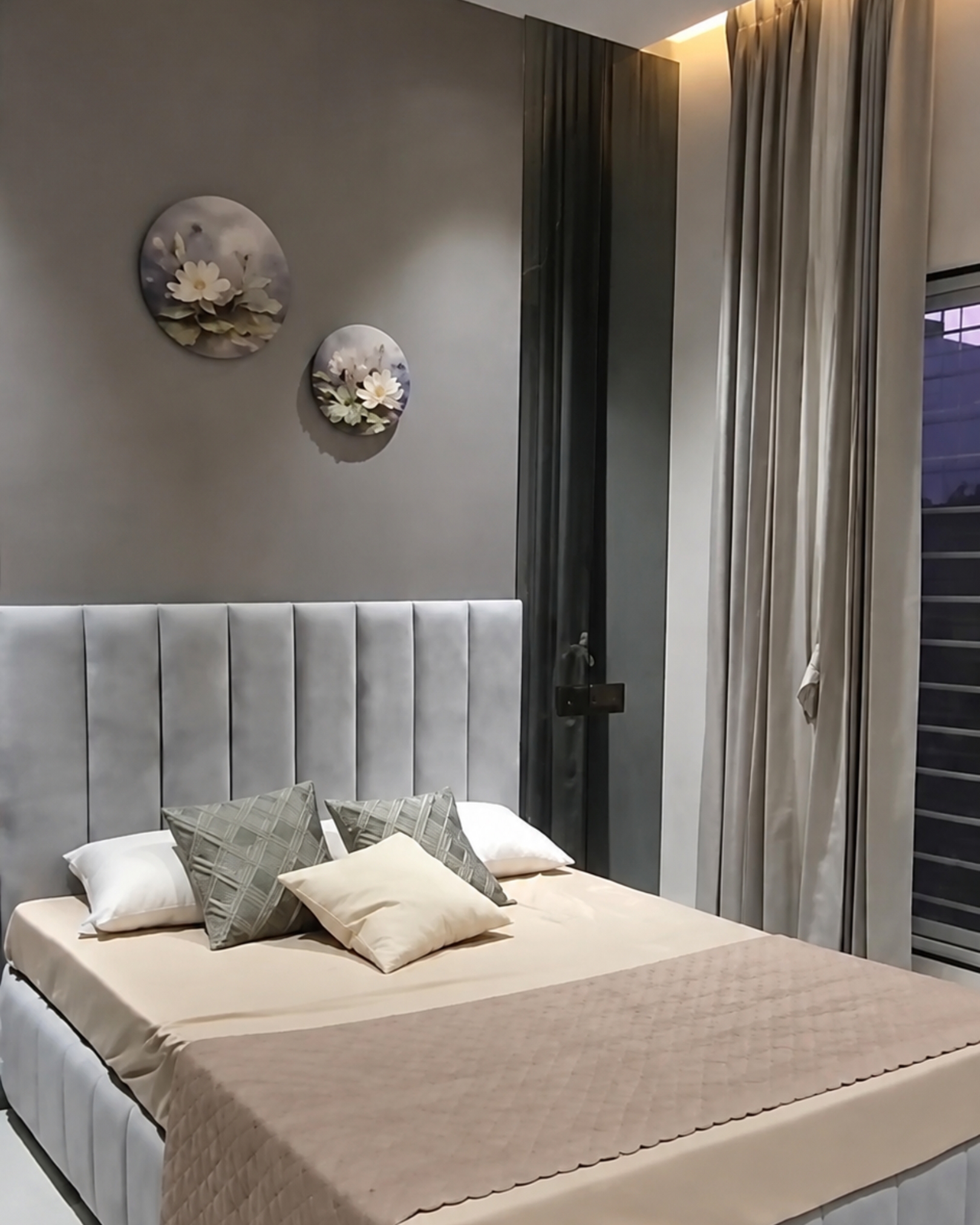 Grey upholstered bed with neutral bedding, patterned cushions and floral wall decorations beside dark fluted panels.