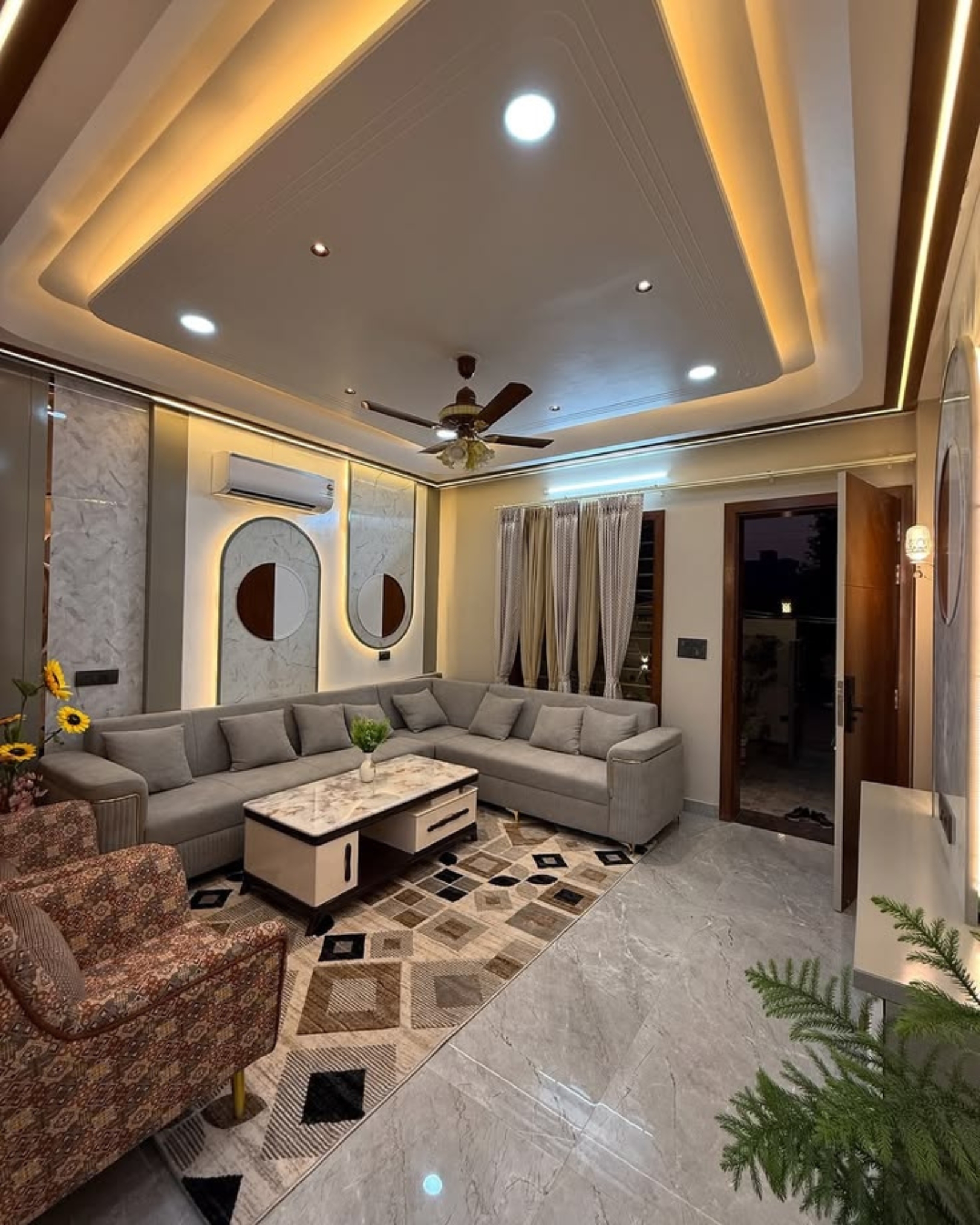 Living room with a grey corner sofa, marble effect wall panels, patterned rug and warm recessed ceiling lighting.