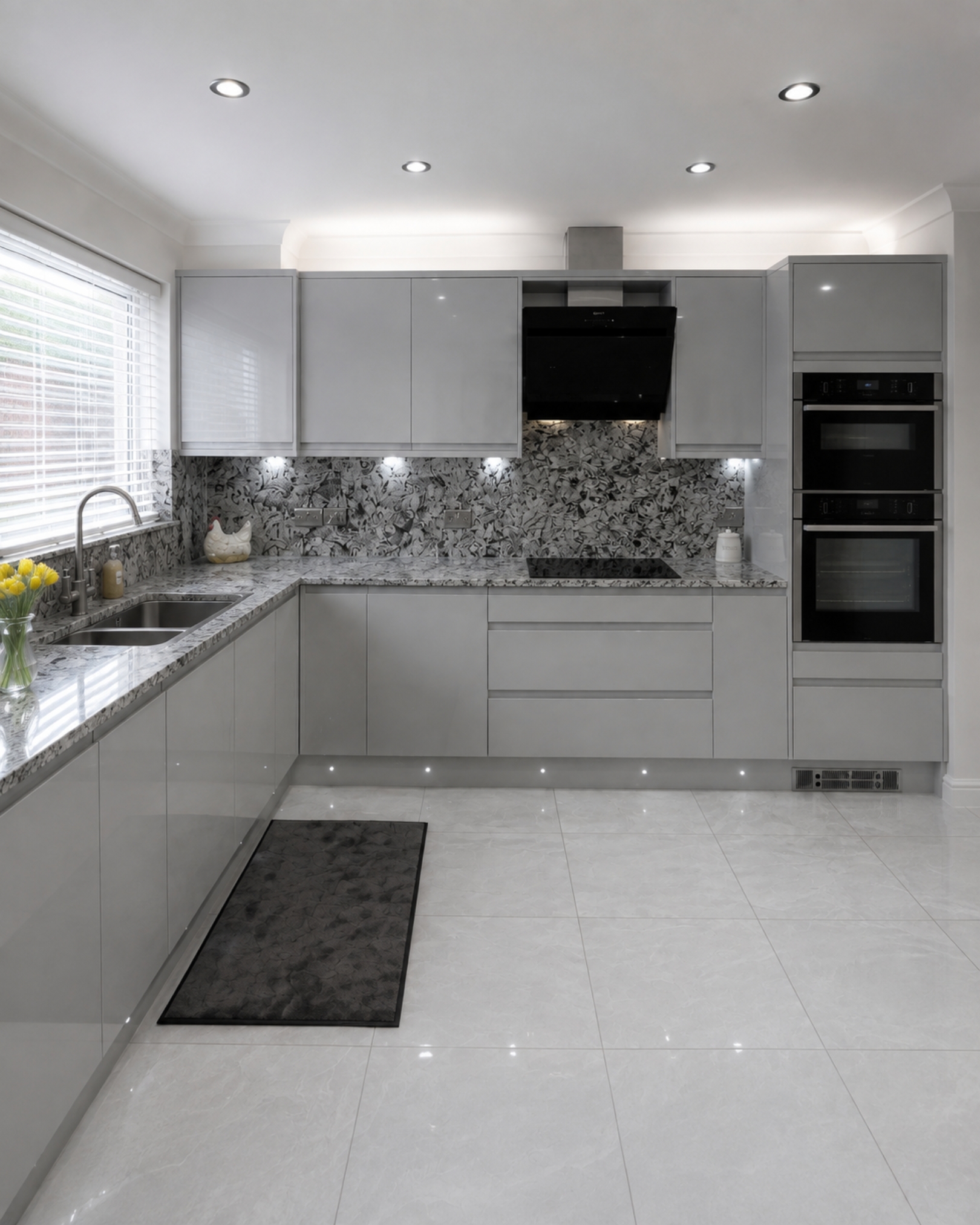 L-shaped kitchen with glossy grey handleless cabinets, patterned countertops and backsplash, stacked built-in ovens, a black extractor hood and under cabinet lighting.